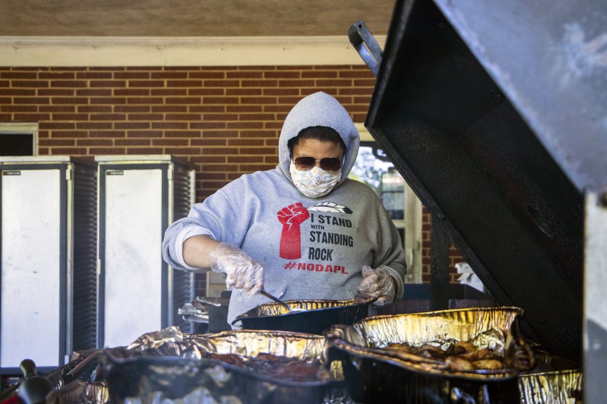 An African American woman wearing a hoodie and mask and sunglasses stands behind a grill holding a large utensil