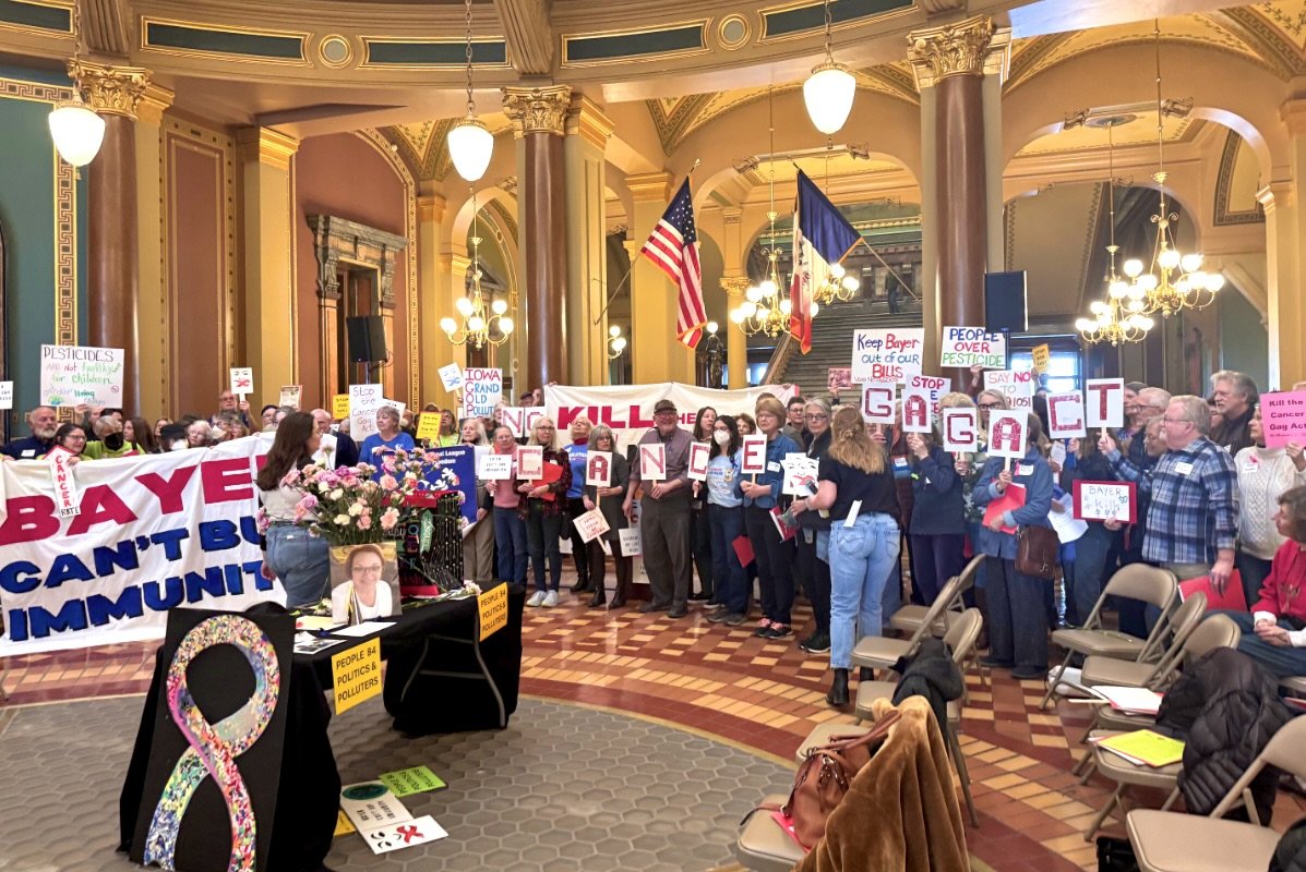 A large group of people hold signs protesting a bill that would stop people from suing Bayer, a maker of pesticides, inside a chamber