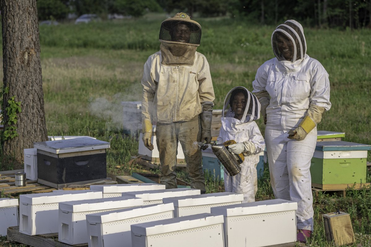 An African American mom, dad, and young son wear white beekkeeping suits and stand in front of honeybee frames for a photo on their farm in North Carolina