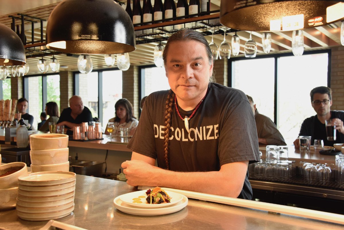 A Native American chef with a long braid leans over a counter with a dish in front of him. He is wearing a t-shirt with the words 